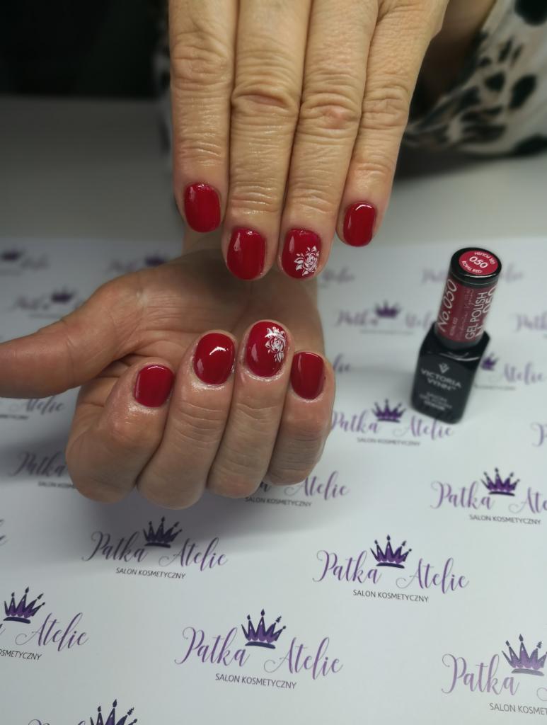 Dłonie z czerwonym manicure hybrydowym, zdobione białymi kwiatowymi wzorami na tle firmowego papieru z logo i buteleczką lakieru.