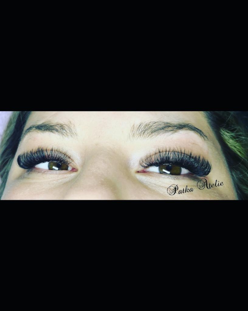 Zbliżenie na oczy kobiety z brązowymi tęczówkami i gęstymi, czarnymi, przedłużonymi rzęsami; widoczny delikatny makijaż brwi i subtelny cień do powiek, w prawym dolnym rogu widoczne logo 'Patka...