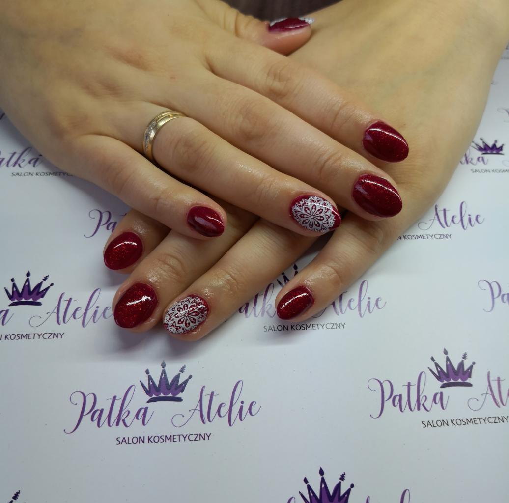 Dłonie z manicure hybrydowym w odcieniach czerwieni z brokatem i wzorem białej rozety na dwóch paznokciach, na tle z logo salonu.