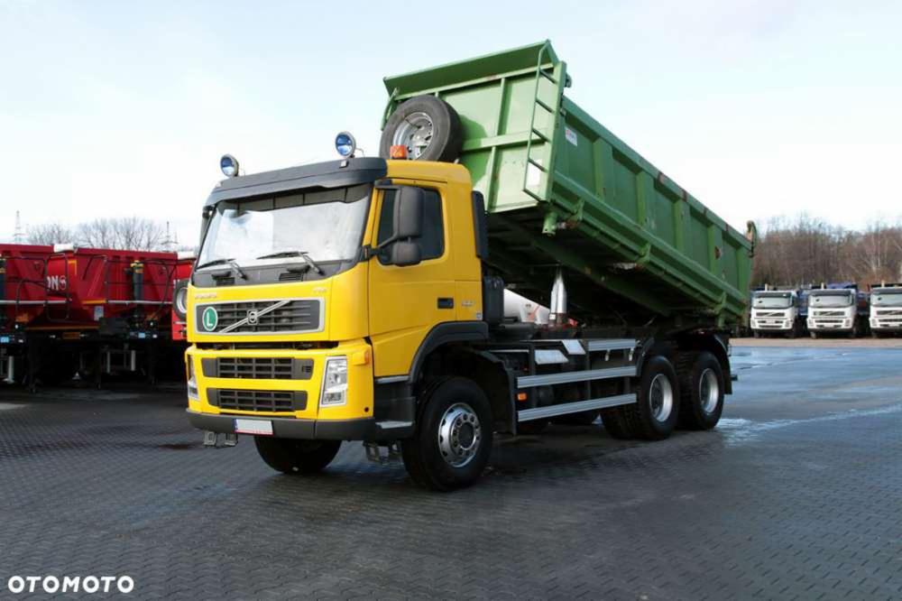 Żółta wywrotka Volvo z podniesioną zieloną skrzynią ładunkową na placu, widoczny zapasowe koło na kabinie i inne ciężarówki w tle.