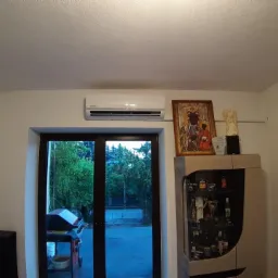 Biały klimatyzator ścienny zamontowany nad drzwiami balkonowymi, obok szafki z alkoholem i obrazem Matki Boskiej Częstochowskiej.