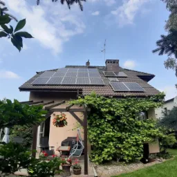 Dom jednorodzinny z brązowym dachem i panelami fotowoltaicznymi, widok z poziomu ogrodu z pergolą obrośniętą bluszczem w pierwszym planie.