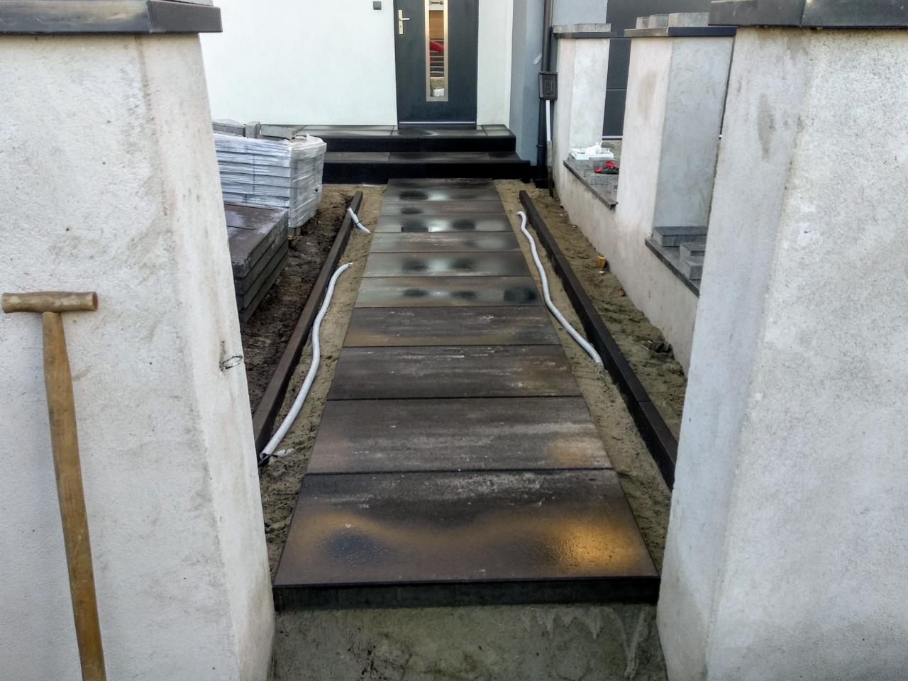 Ścieżka z ciemnych płyt betonowych w trakcie układania, widoczne białe rury i paleta z kostką, ograniczona betonowymi słupkami
