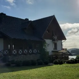 Dwupiętrowy budynek z ciemnym dachem, drewnianymi elementami i zielonym trawnikiem przed nim, w tle jezioro i las pod pochmurnym niebem.