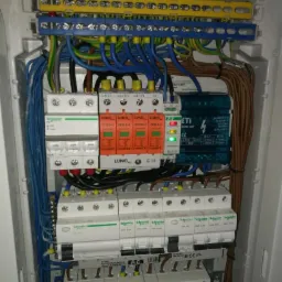 Otwarta skrzynka elektryczna z widocznymi bezpiecznikami, modułami przepięciowymi LUNO i ETI, listwami zaciskowymi, oraz licznymi przewodami w różnych kolorach, ułożonymi wewnątrz obudowy.