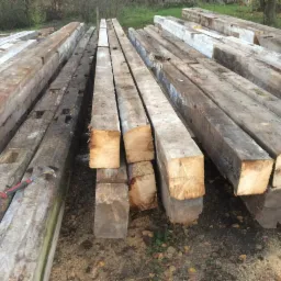 Stare, drewniane belki o kwadratowym przekroju, ułożone w stos na ziemi, z widocznym młotkiem ciesielskim obok jednej z belek.