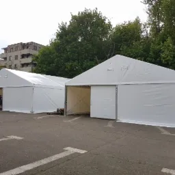 Dwa białe namioty magazynowe z metalową konstrukcją na betonowym placu, z widocznymi paletami i osobami w tle.