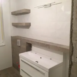 Nowoczesna łazienka w trakcie wykończenia: biała umywalka z szufladami, kamienne półki i blat, surowe ściany w tle.