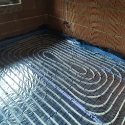 Montaż ogrzewania podłogowego w surowym wnętrzu, widoczne rury grzewcze marki Keller zamocowane na folii z siatką, ściany z czerwonej cegły.