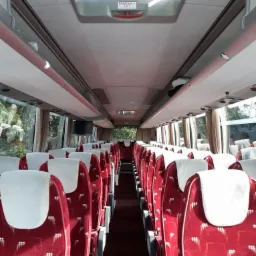 Wnętrze autobusu turystycznego z czerwonymi fotelami z białymi zagłówkami i widocznym znakiem wyjścia awaryjnego.