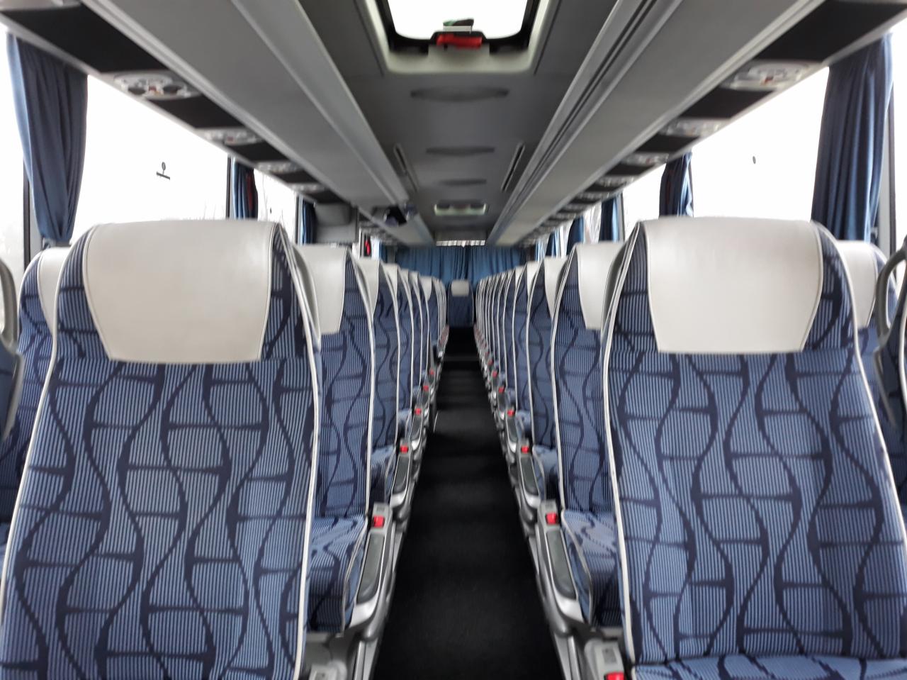 Wnętrze autobusu z rzędami niebieskich foteli z geometrycznym wzorem i białymi zagłówkami, widok z perspektywy pasażera.