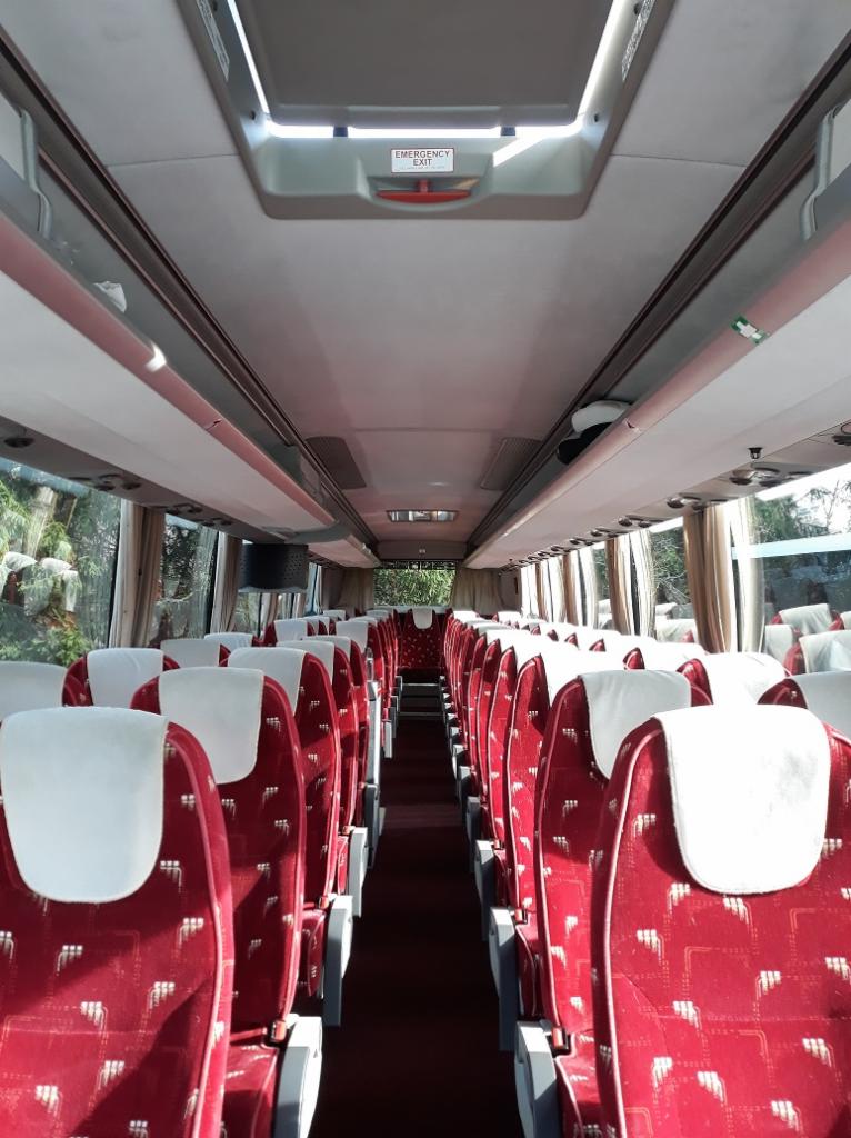 Wnętrze autobusu turystycznego z czerwonymi fotelami z białymi zagłówkami i widocznym znakiem wyjścia awaryjnego.