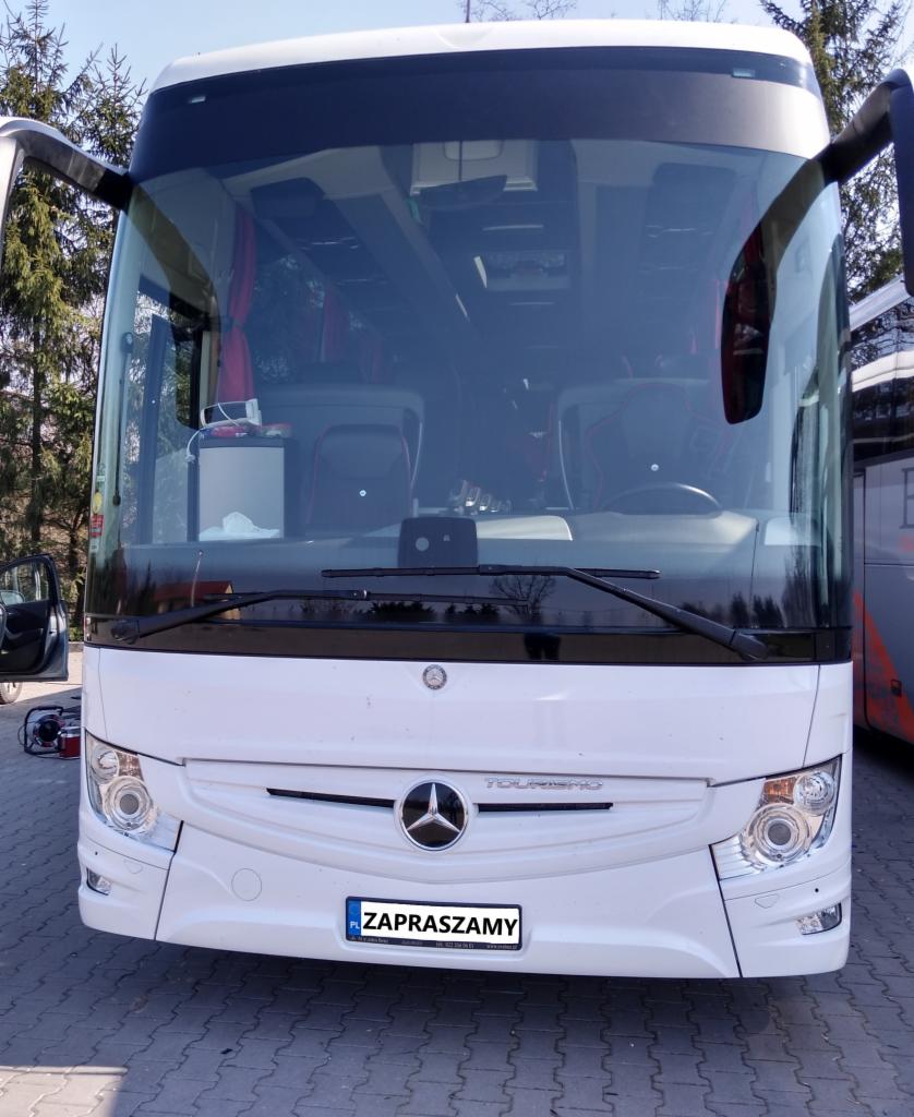 Przód białego autobusu turystycznego marki Mercedes z otwartymi drzwiami, widoczne drzewa w tle, tablica rejestracyjna z napisem 'Zapraszamy'.
