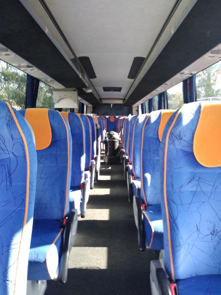 Wnętrze autobusu turystycznego z rzędami niebieskich foteli z pomarańczowymi zagłówkami, widok z przodu kabiny pasażerskiej, czarna wykładzina podłogowa i metalowy kosz na śmieci na końcu przejścia.