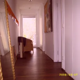 Korytarz z ciemnymi panelami podłogowymi, widok na lustro w złotej ramie, drzwi balkonowe i wiklinowy fotel.