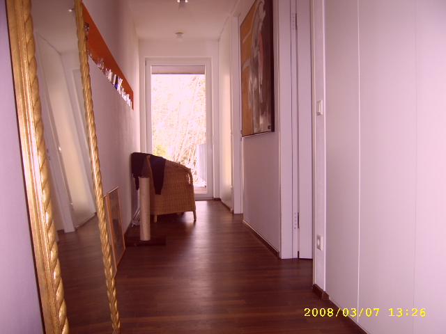 Korytarz z ciemnymi panelami podłogowymi, widok na lustro w złotej ramie, drzwi balkonowe i wiklinowy fotel.