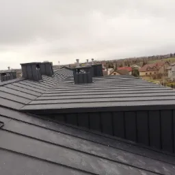 Nowoczesny, grafitowy dach z blachy na rąbek stojący z licznymi kominami wentylacyjnymi na tle zabudowy jednorodzinnej i pochmurnego nieba.