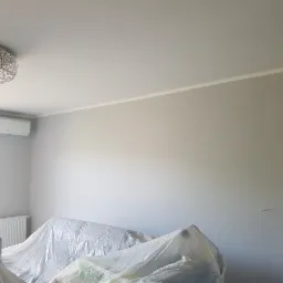 Pokój w Bielczy przygotowany do malowania: meble zabezpieczone folią, ściany świeżo pomalowane na szaro, widoczny klimatyzator i kryształowy żyrandol.