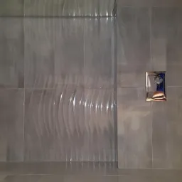 Szare płytki ceramiczne z delikatnym, falistym wzorem na ścianie kabiny prysznicowej, widoczny chromowany przycisk spłuczki oraz fragment toalety.