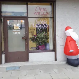 Fasada salonu z brązowymi drzwiami i oknem, logo Wella i Isabelle Lancray, godziny otwarcia na drzwiach, świąteczna dekoracja w oknie i dmuchany Święty Mikołaj przed wejściem.