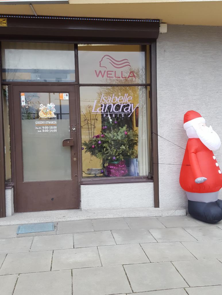 Fasada salonu z brązowymi drzwiami i oknem, logo Wella i Isabelle Lancray, godziny otwarcia na drzwiach, świąteczna dekoracja w oknie i dmuchany Święty Mikołaj przed wejściem.