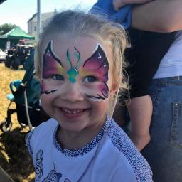 Ascom - Uśmiechnięta dziewczynka z pomalowaną twarzą w motyla, na tle plenerowej imprezy z wózkiem dziecięcym i budynkiem w oddali.