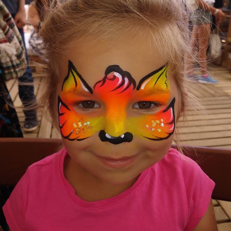 Uśmiechnięta dziewczynka z pomalowaną twarzą w motywy zwierzęce: pomarańczowo-żółty pyszczek z czarnym nosem i uszami, dodatkowo ozdobiony brokatem.