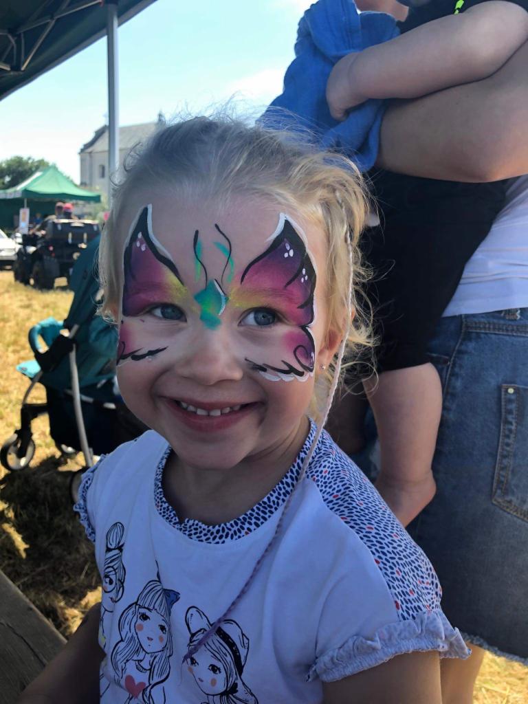 Uśmiechnięta dziewczynka z pomalowaną twarzą w motyla, na tle plenerowej imprezy z wózkiem dziecięcym i budynkiem w oddali.