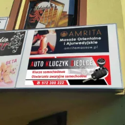 Zewnętrzny szyld reklamowy z ofertą usług: dorabianie kluczy samochodowych i awaryjne otwieranie aut, umieszczony nad salonem fryzjerskim i gabinetem masażu.