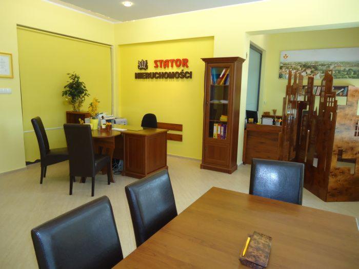 Jasne biuro agencji nieruchomości z drewnianymi meblami, logo firmy na ścianie i dekoracyjnym parawanem z motywem miejskim.