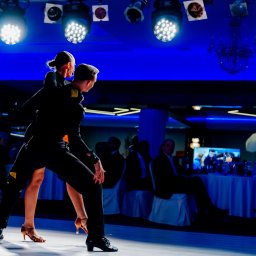 Leszek Dopke Fotografia - Para tancerzy w czarnych strojach prezentuje dynamiczną pozę na parkiecie oświetlonym reflektorami, w tle widoczni widzowie przy stolikach.