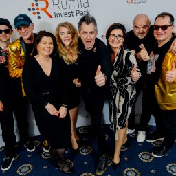 Leszek Dopke Fotografia - Grupa osób, w tym mężczyźni w złotych marynarkach, pozuje na tle ściany z logo Rumia Invest Park, wszyscy pokazują kciuki w górę.
