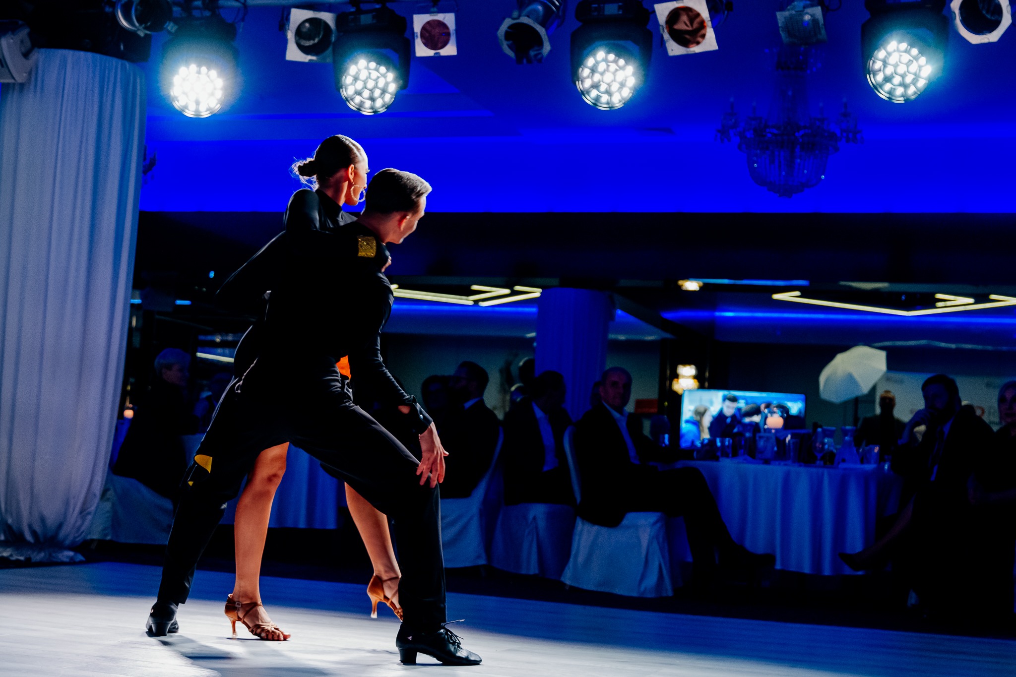 Para tancerzy w czarnych strojach prezentuje dynamiczną pozę na parkiecie oświetlonym reflektorami, w tle widoczni widzowie przy stolikach.