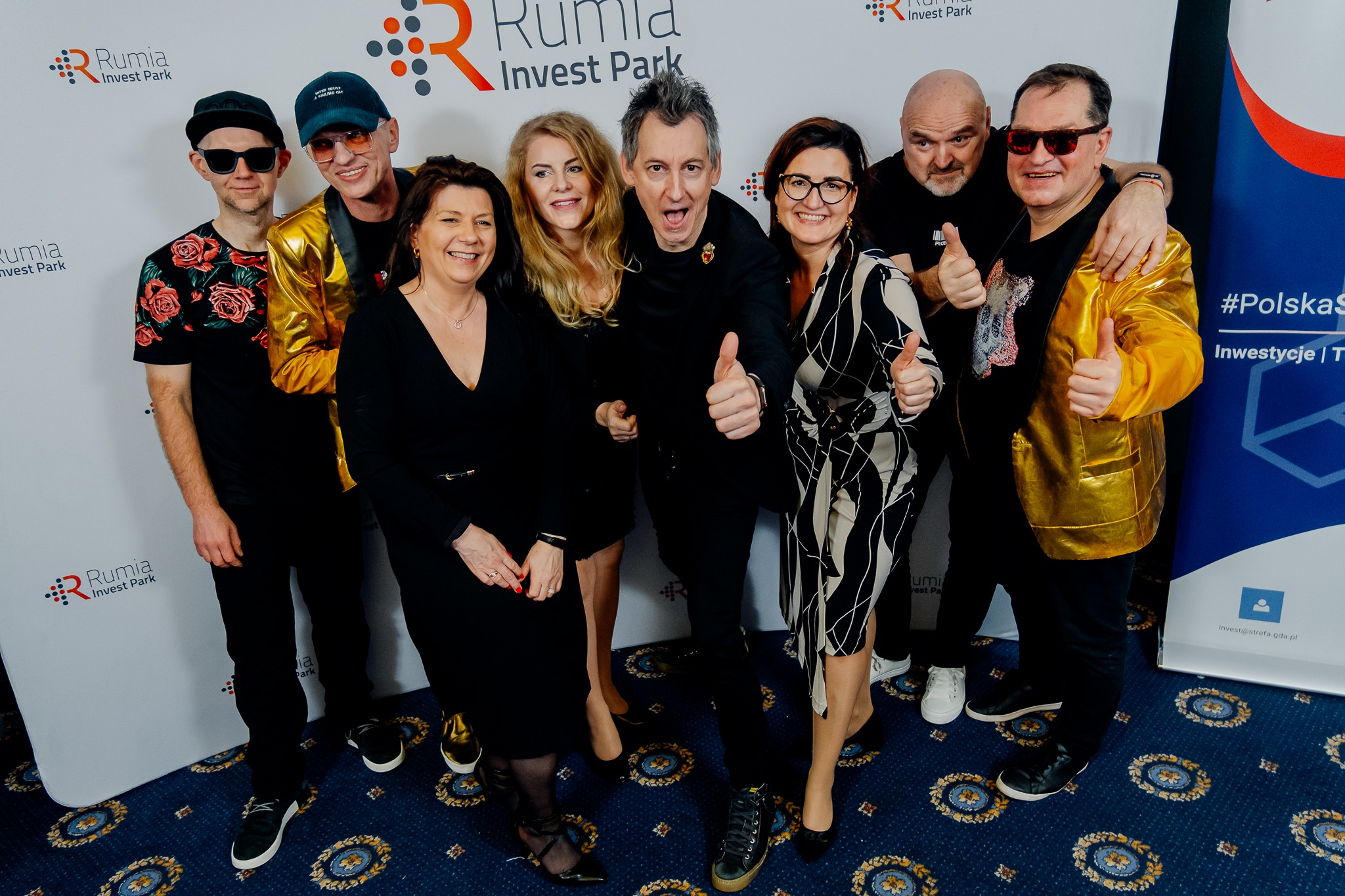 Grupa osób, w tym mężczyźni w złotych marynarkach, pozuje na tle ściany z logo Rumia Invest Park, wszyscy pokazują kciuki w górę.