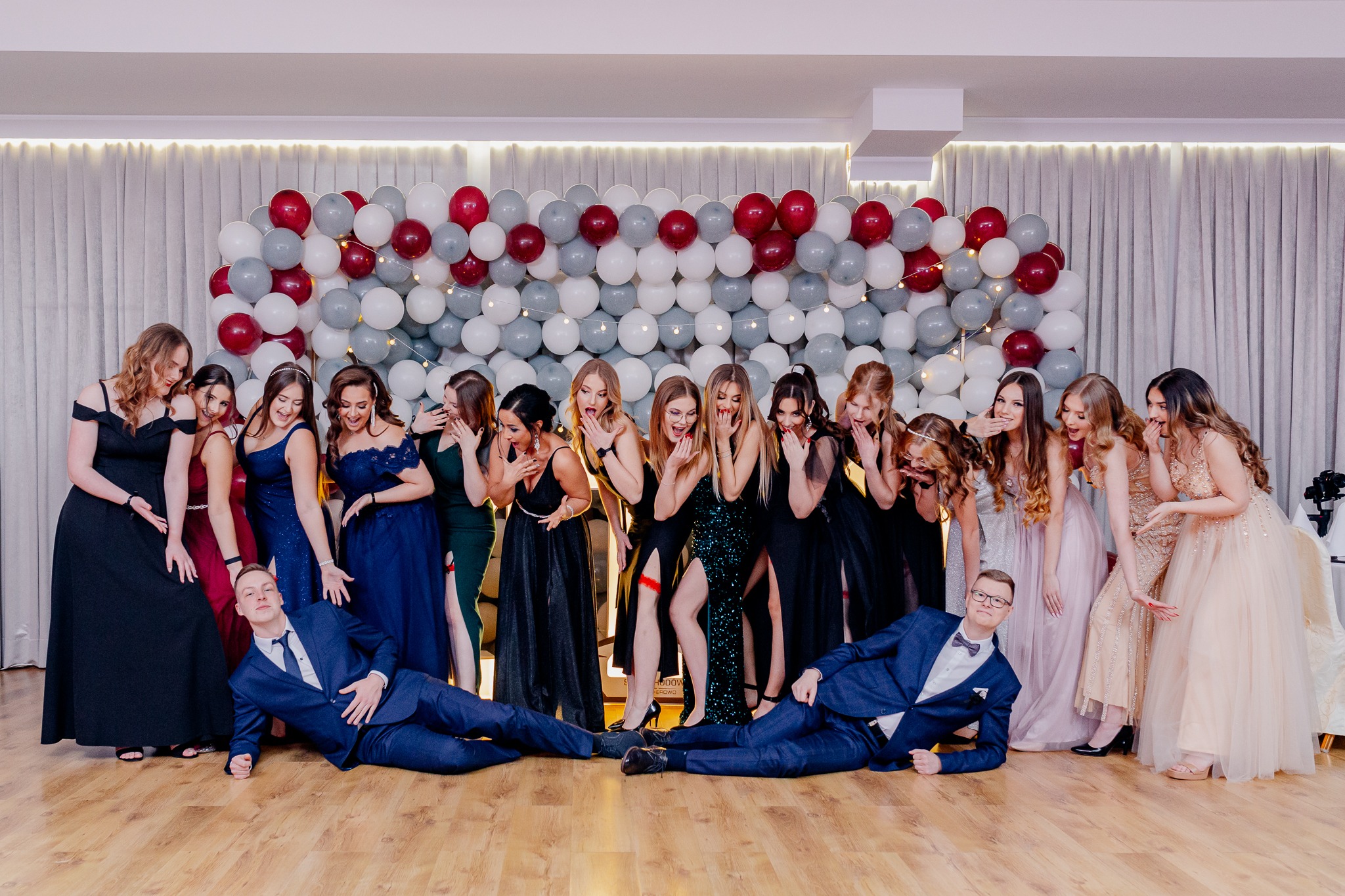 Grupa elegancko ubranych kobiet i dwóch mężczyzn w garniturach pozuje na tle ściany z balonów w Orle, z zaskoczonymi minami i rozbawieniem.