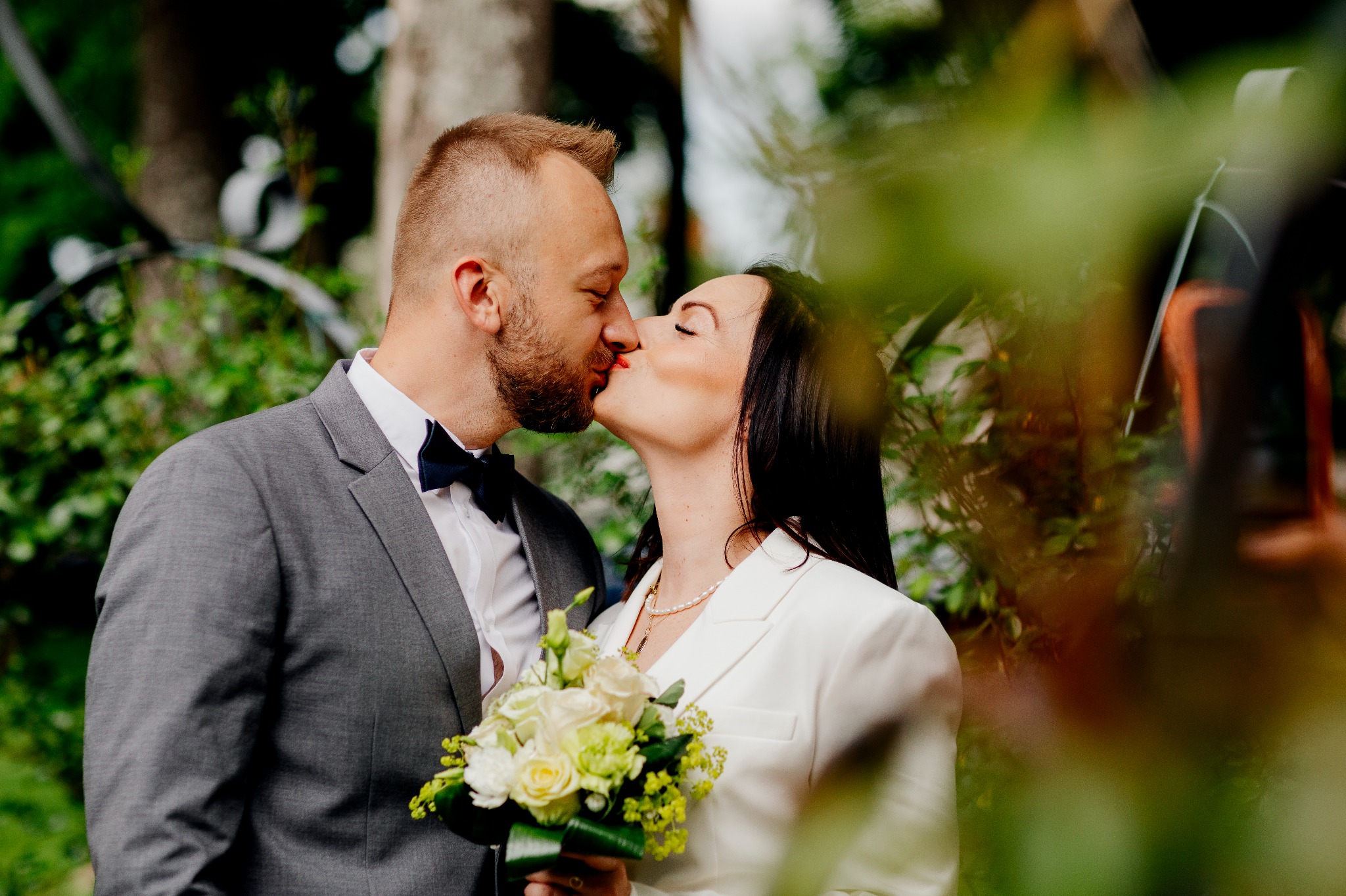 Pocałunek młodej pary: mężczyzna w szarym garniturze i muszce oraz kobieta w białym żakiecie trzymająca bukiet z białych i żółtych róż.