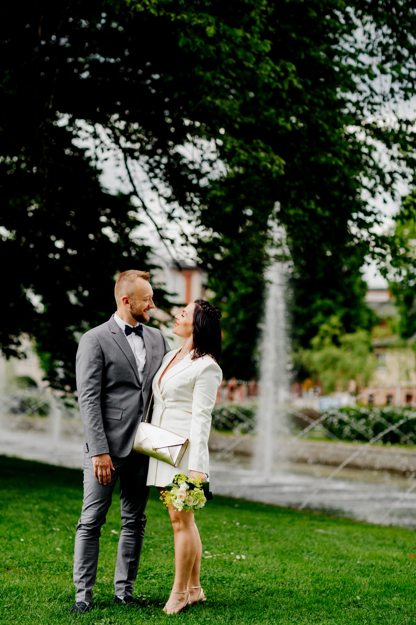 Uśmiechnięta para młoda w parku na tle fontanny, panna młoda w białym garniturze trzyma bukiet, pan młody w szarym garniturze z muchą.