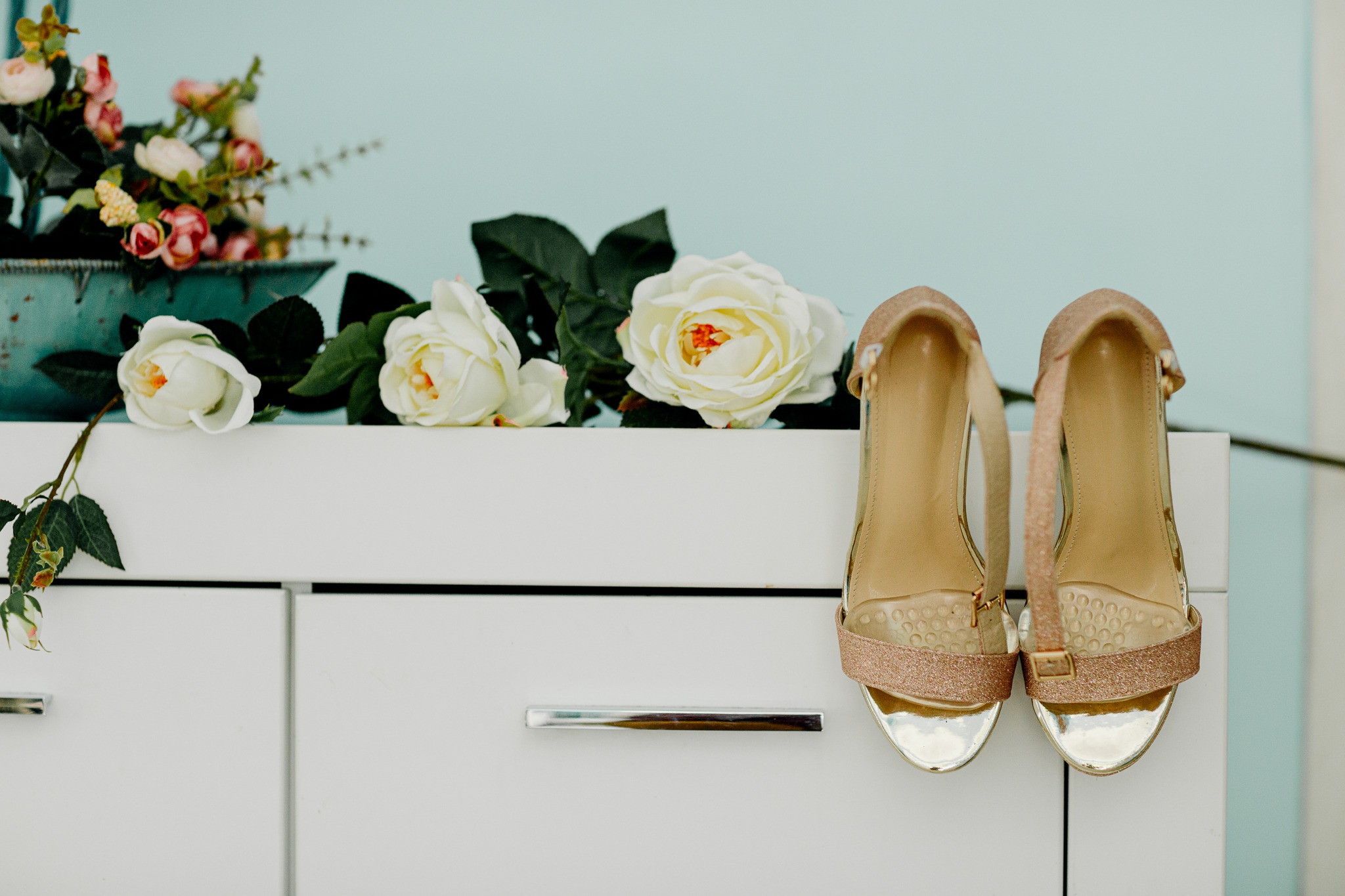 Para złotych butów na obcasie z brokatowymi paskami stoi obok białych róż na białej komodzie, na tle jasnoniebieskiej ściany.