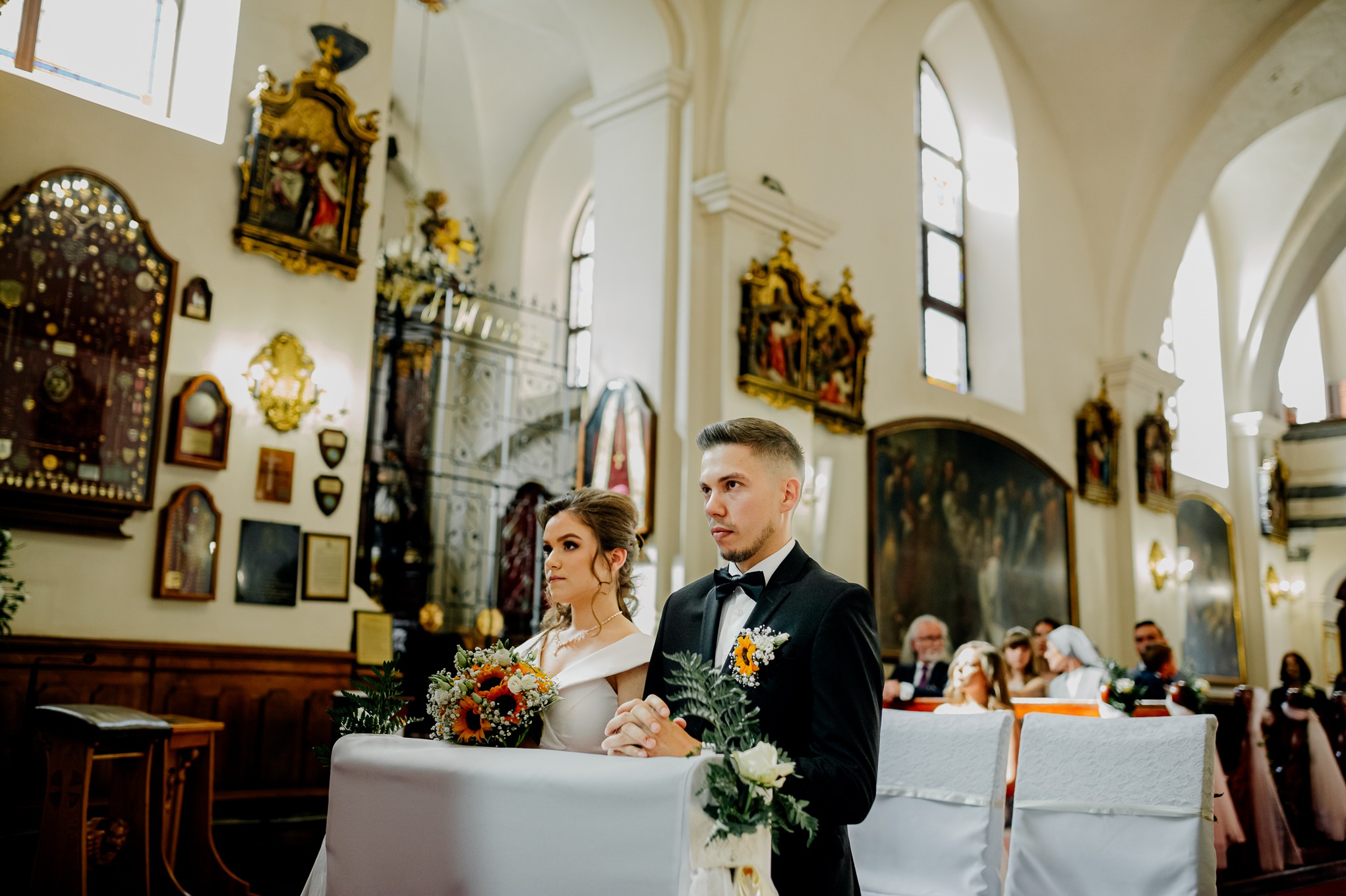Para młoda w kościele podczas ceremonii ślubnej, panna młoda z bukietem słoneczników, pan młody w czarnym smokingu z butonierką, zdobione ściany kościoła w tle.