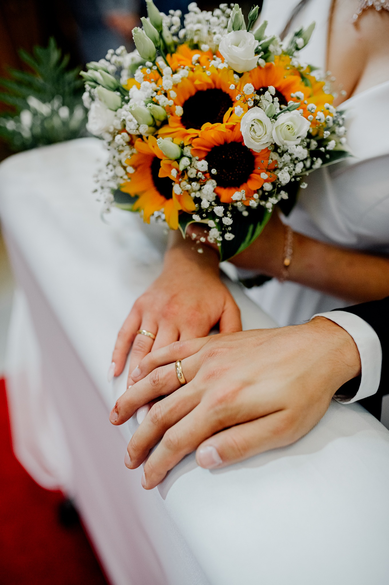 Dłonie nowożeńców ze złotymi obrączkami spoczywają na białym obrusie obok bukietu ślubnego z białych róż i słoneczników.