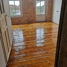 Odnowiona drewniana podłoga w pokoju z odsłoniętą cegłą na ścianie, widok przez otwarte drzwi.