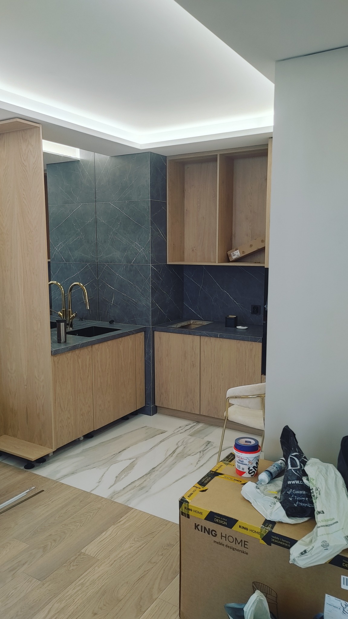 Fragment kuchni w trakcie wykańczania: widoczne drewniane szafki, czarny blat i płytki imitujące marmur na ścianie, złota armatura, karton po meblach i pojemnik z gładzią na podłodze.