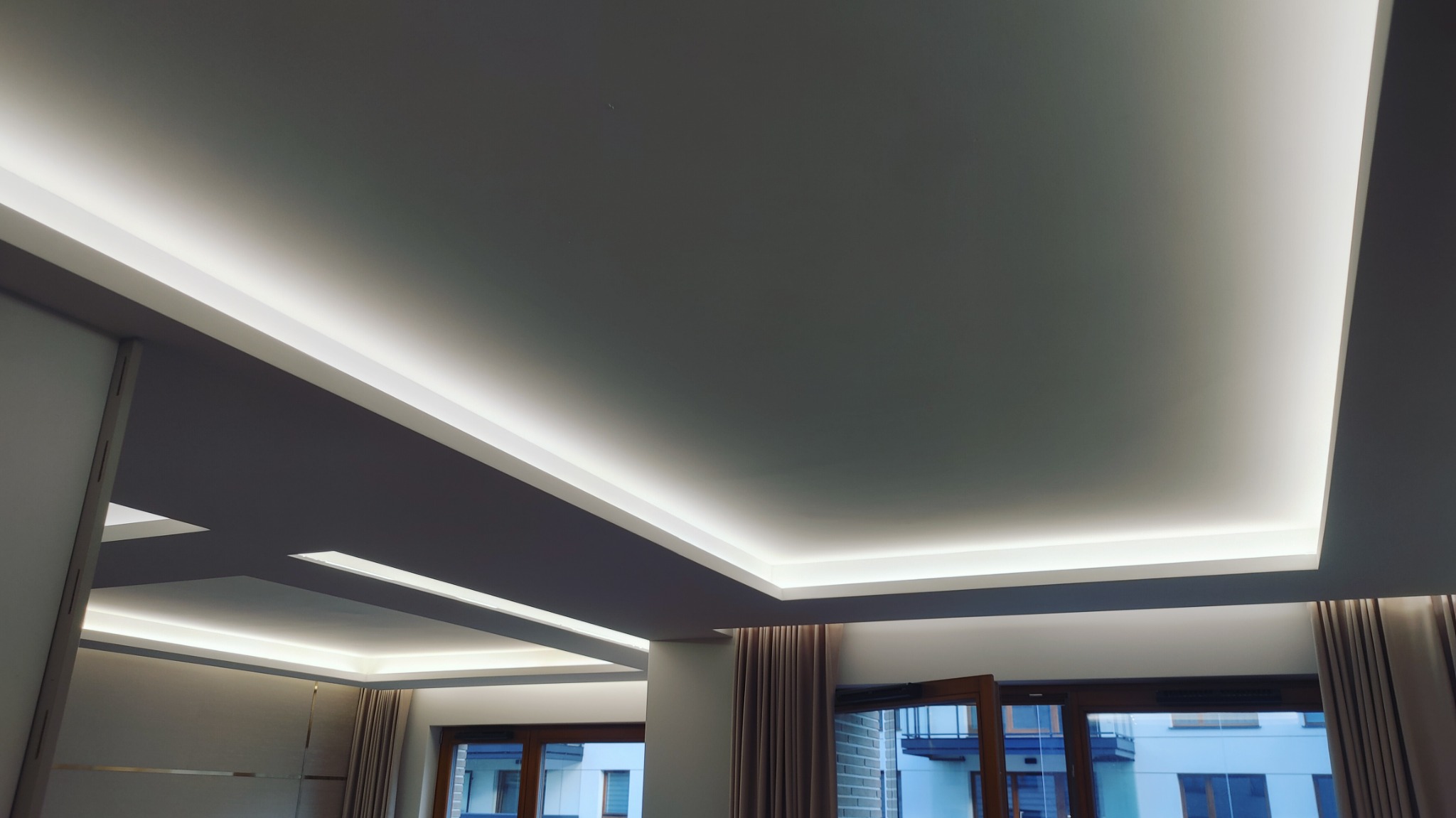 Nowoczesny sufit podwieszany z oświetleniem LED w jasnym wnętrzu, widok z dołu, okna z zasłonami w tle.