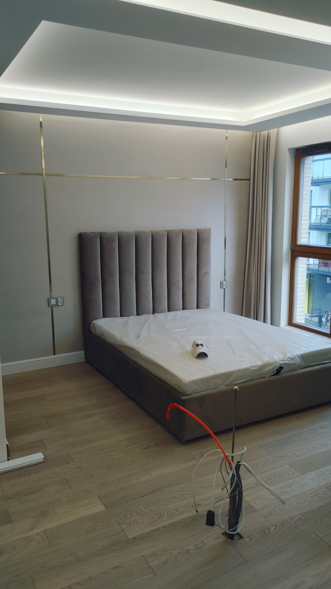 Sypialnia w trakcie wykańczania: łóżko z szarym wezgłowiem, podłoga z jasnego drewna, widoczne kable wystające z podłogi, listwy dekoracyjne na ścianie.