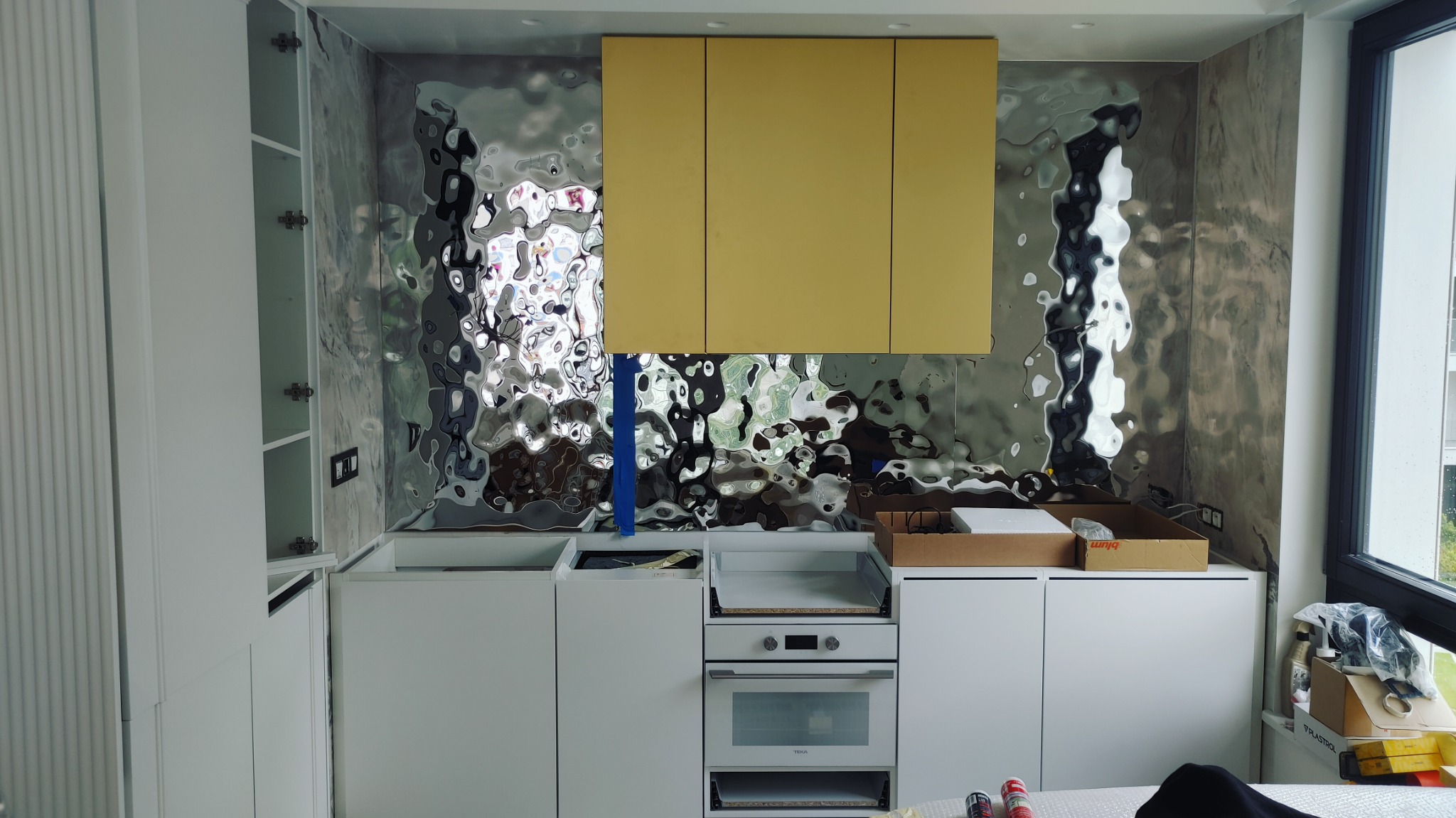Montaż nowoczesnej kuchni z frontami w kolorze białym i żółtym, z dekoracyjnym panelem ściennym o fakturze przypominającej płynny metal.