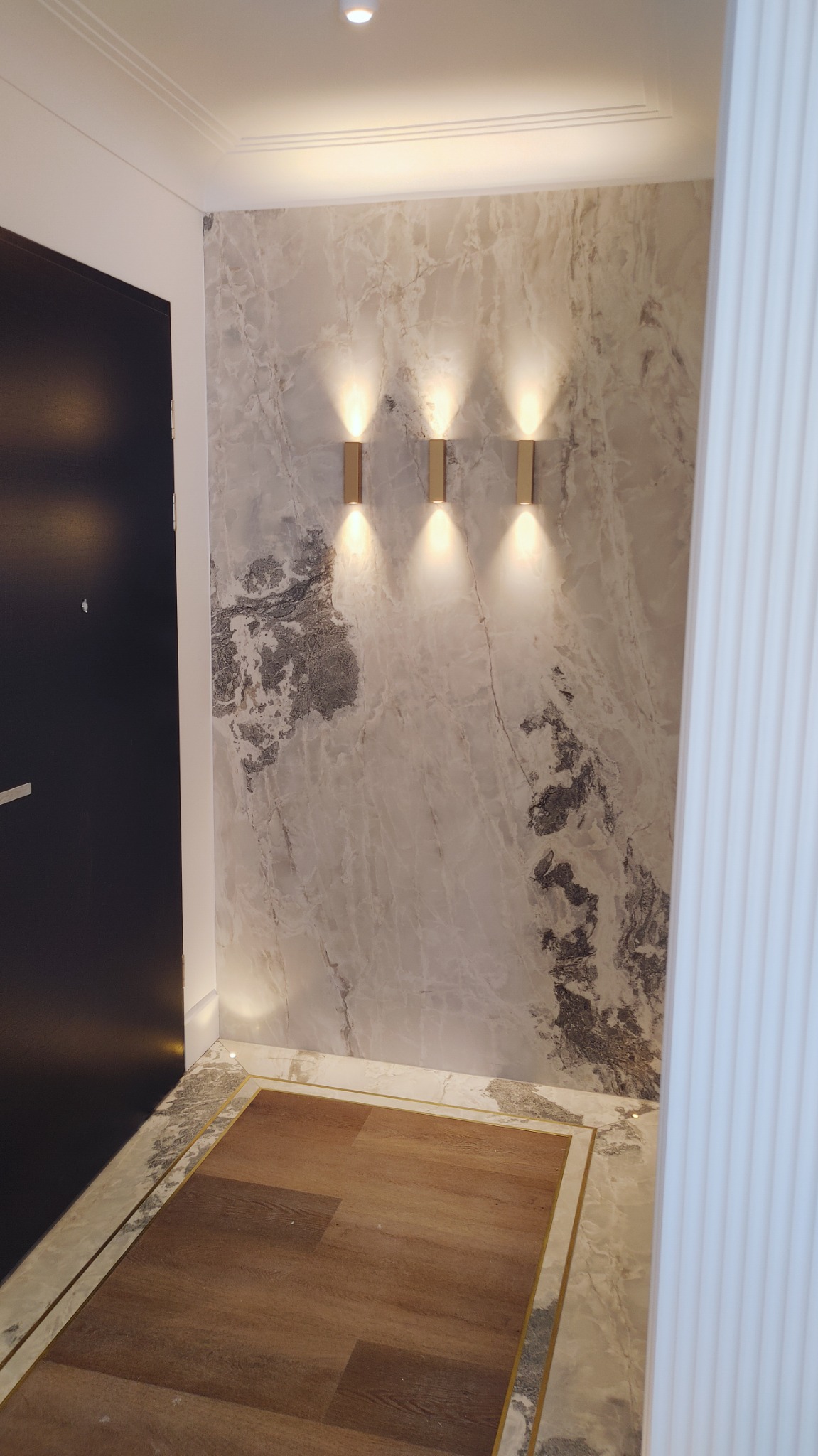Nowoczesne wejście: ciemne drzwi, marmurowa ściana z trzema złotymi kinkietami, drewniana podłoga w marmurowej ramie, oświetlenie sufitowe.