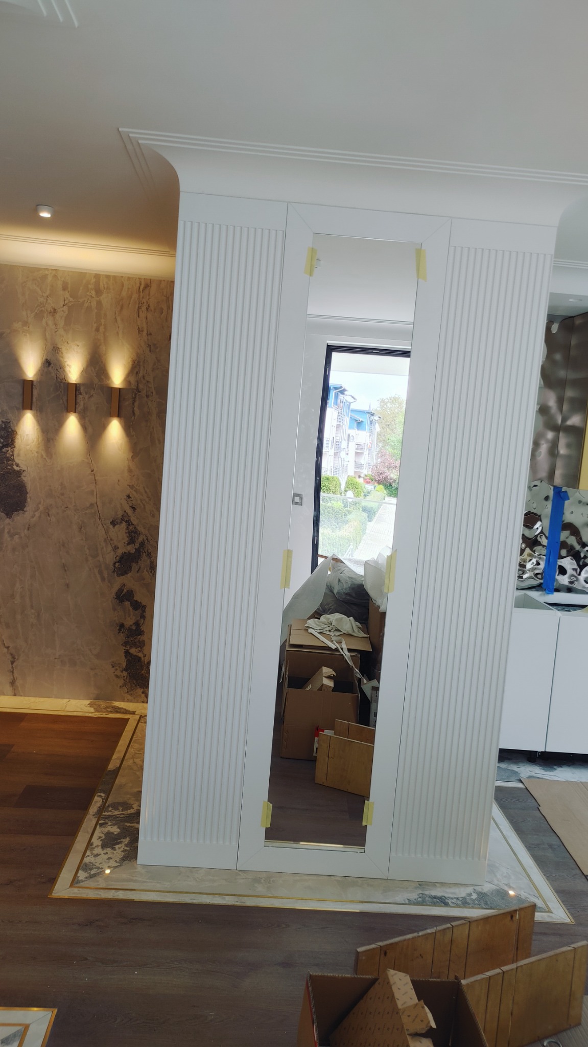 Montaż lustrzanych drzwi w zabudowie z białymi, frezowanymi panelami ściennymi, zabezpieczonych taśmą malarską, widoczne kartony i materiały budowlane w tle, marmurowa ściana z oświetleniem.