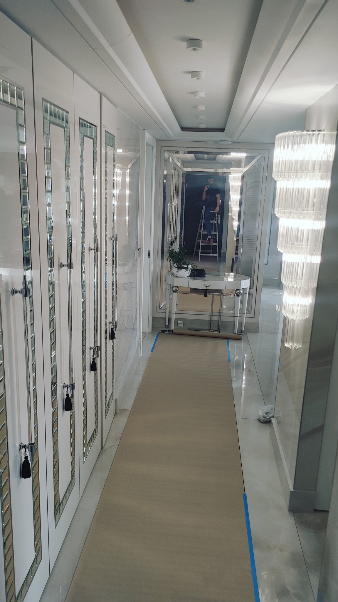 Lustrzane szafy wnękowe w długim korytarzu, zabezpieczona podłoga, pracownik na drabinie widoczny w lustrze, elegancki żyrandol.