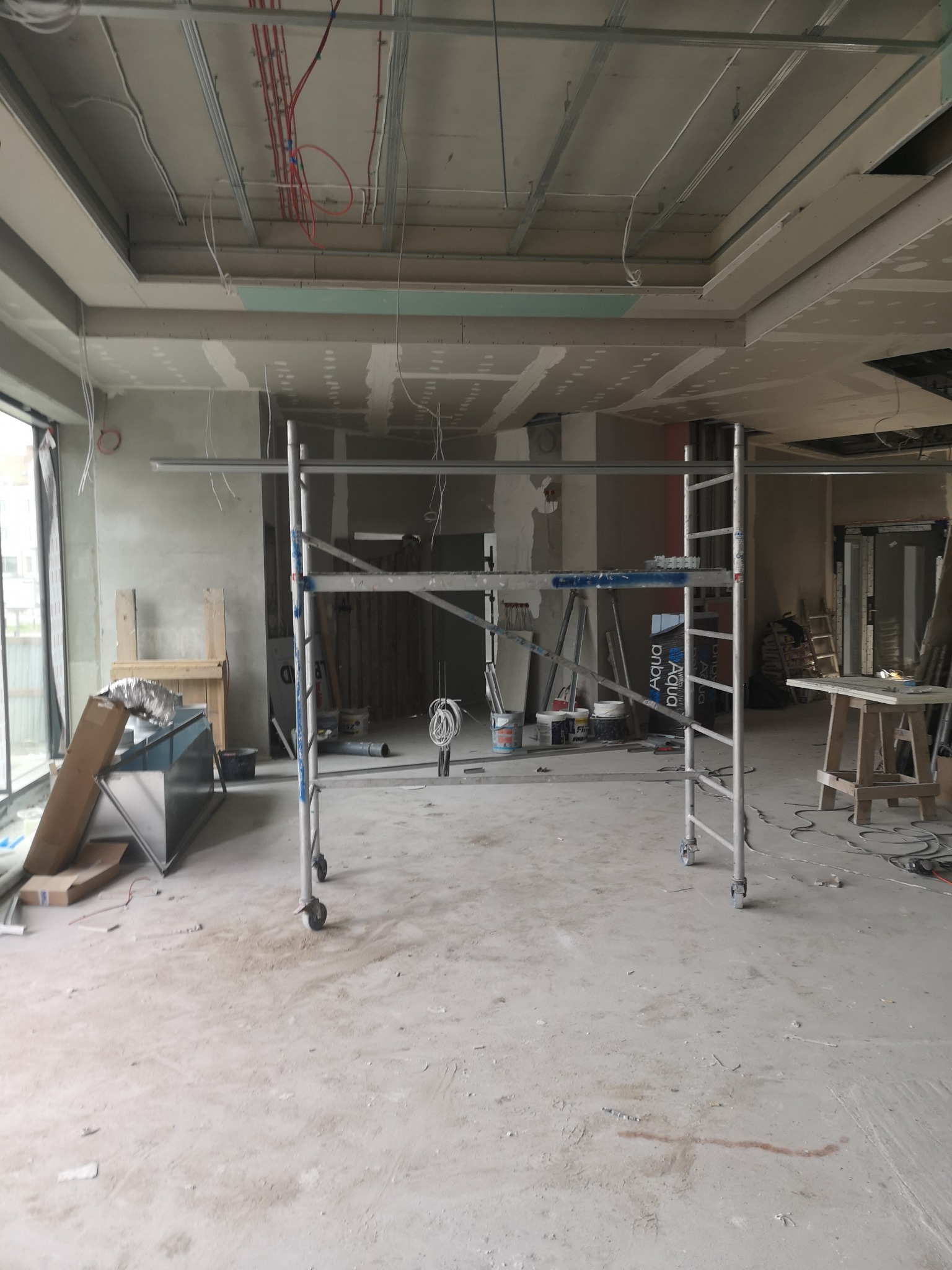 Wnętrze w trakcie remontu: widoczne rusztowanie, częściowo wykończony sufit z płyt kartonowo-gipsowych, instalacje elektryczne pod sufitem oraz materiały budowlane na podłodze.