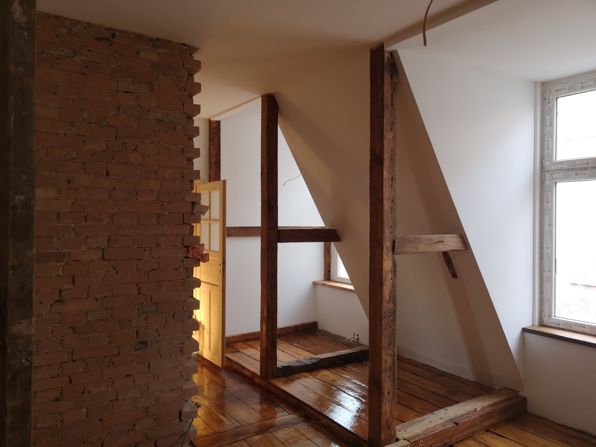 Pokój na poddaszu w trakcie remontu: fragment ściany z cegły, drewniana konstrukcja nośna, surowa podłoga z desek, okno z PCV.